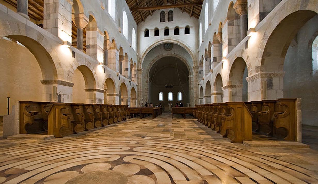 A large church lined with carved archways and ornate floor patterning carved wooden benches for seating reach far into the back where a mass is being held